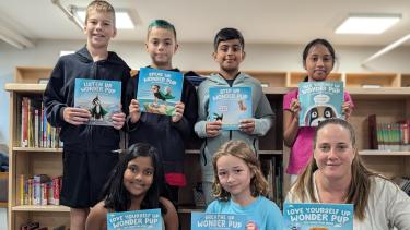 Six students and 1 Adult holding up books in front of them in the library.